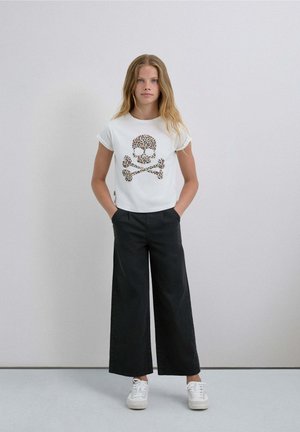 Camiseta blanca con una gráfica de calavera y huesos cruzados con estampado de leopardo, combinada con pantalones negros de pierna ancha y zapatillas blancas.