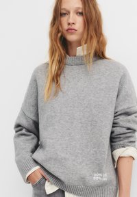 Maglione grigio lavorato a maglia con colletto alto, orlo e dettagli delle maniche a coste. Presenta una scritta sul davanti: "50% idt 50% ide."