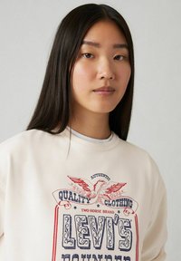 Felpa crema con grafica in stile vintage che presenta un design con testo rosso e blu e un'aquila. Tessuto morbido, vestibilità rilassata, scollatura a girocollo.