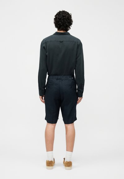 Homme aux cheveux bouclés et foncés portant une chemise à manches longues foncée, un short bleu marine, des chaussettes blanches et des chaussures marron, debout de dos.