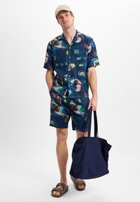 Marin blauw shirt met korte mouwen en bijpassende shorts met abstracte kleurrijke patronen, gecombineerd met bruine sandalen en een marineblauwe tote bag.