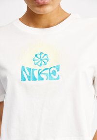 Valkoinen puuvillapaita, jossa on sininen brodeerattu logo, jossa on kukka-aihe sanan "NIKE" yläpuolella. Lyhyet hihat ja pyöreä kaula-aukko.