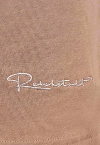 Tessuto a maglia beige con una firma ricamata in evidenza in filo bianco. La texture è liscia con una leggera elasticità.