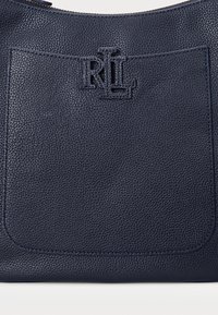 Marineblaue Handtasche aus Leder mit strukturiertem Finish, ausgestattet mit einem auffälligen geprägten Logo und einer großen Fronttasche zur Aufbewahrung.