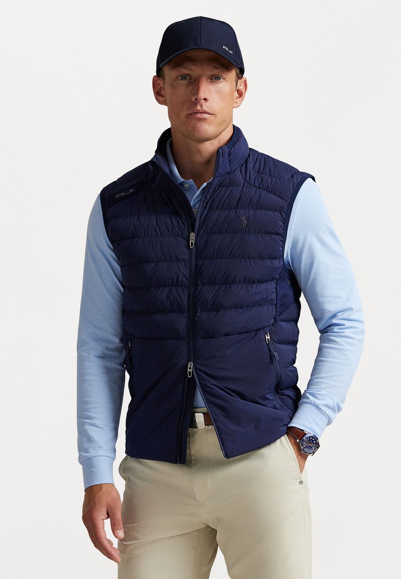 Navy puffer vest with quilted design, side pockets, and zip closure, worn over a light blue long-sleeve shirt. Detailed with a logo.