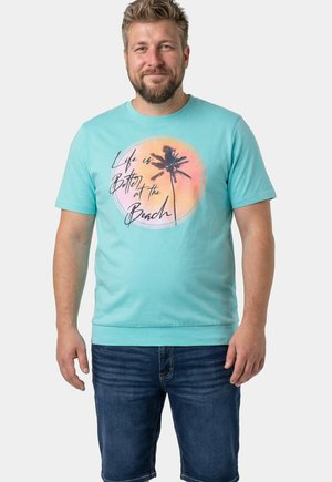 Bärtiger Mann trägt ein türkisfarbenes T-Shirt mit Palmen- und Sonnenuntergangsgrafik und dem Text "Life is Better at the Beach", kombiniert mit Jeansshorts.