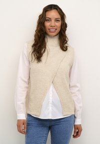 Pull à col roulé sans manches en tricot beige avec un design croisé, porté par-dessus une chemise blanche à col, associé à un jean bleu.