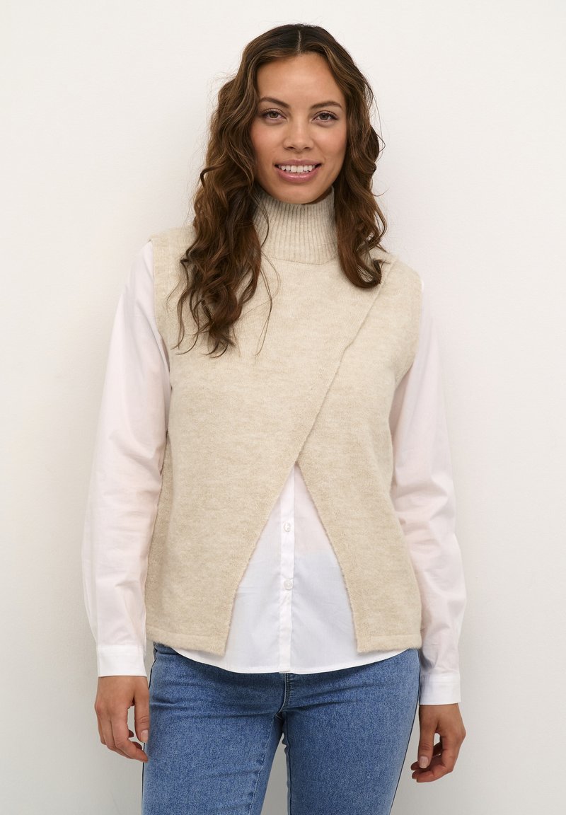 Pull à col roulé sans manches en tricot beige avec un design croisé, porté par-dessus une chemise blanche à col, associé à un jean bleu.