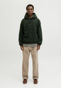 Sweat à capuche en polaire vert forêt avec des poignets élastiques, superposé sur une chemise à carreaux, associé à un pantalon beige et des chaussures marron.