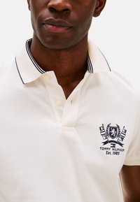 Camisa polo crema hecha de tejido texturizado, con un cuello con ribetes negros, tapeta de tres botones y logotipo bordado con elementos decorativos.