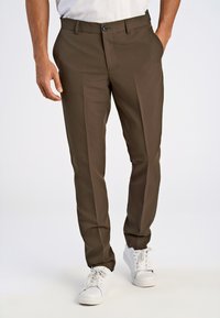 Mann in braunen maßgeschneiderten Hosen und weißen Sneakers, der mit einer Hand in der Tasche vor einem schlichten Hintergrund steht.