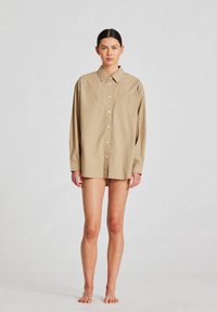 Beige oversized gombos ing egyetlen mellzsebbel. Hosszú ujjú, puha tapintású és laza fazonú. A modell mezítláb áll.