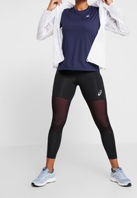 Completo sportivo con maglietta a maniche corte blu navy, leggings neri con dettagli in rete e scarpe da corsa azzurro chiaro. Giacca leggera bianca.