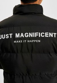Jachetă neagră căptușită cu guler înalt, având pe spate text alb: "DOAR MAGNIFIC" deasupra "FĂ-L SĂ SE ÎNTÂMPLCE." Textură netedă.