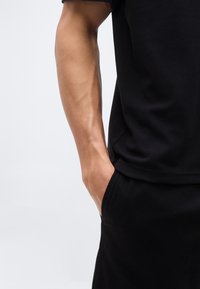 Δεξί χέρι και χέρι ακουμπισμένο στην τσέπη, φορώντας μαύρο πουκάμισο με κοντά μανίκια και μαύρο παντελόνι, σε απλό φωτεινό φόντο.