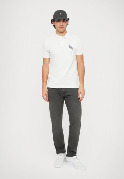 Camisa polo branca com um detalhe bordado em azul e laranja, combinada com calças cinza-escuras e sapatilhas brancas, acompanhada por um boné preto.