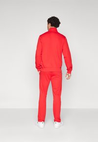 Chándal deportivo rojo con cuello alto, puños acanalados y pantalones a juego. La tela parece suave y elástica. Combinado con zapatillas blancas.