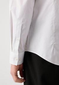 Camisa de manga larga con rayas blancas y puño abotonado. Presenta un dobladillo curvado y una textura suave. Mano parcialmente visible en el lado.