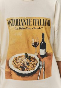 Crèmekleurig t-shirt met een kleurrijke illustratie van pasta, een wijnglas en een fles. De tekst luidt "Ristorante Italiano" en "La Dolce Vita, a Tavola."