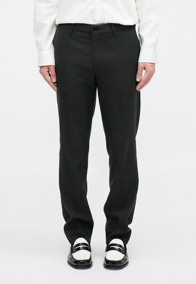 Pantaloni neri su misura con taglio dritto, chiusura con cerniera e bottone, abbinati a mocassini bianco e nero e a una camicia bianca a maniche lunghe.