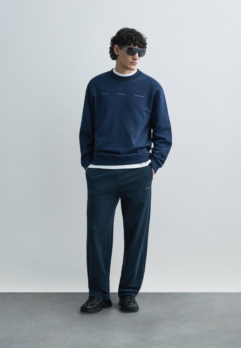 Sweat-shirt bleu marine avec texte brodé, porté sur une chemise blanche. Pantalon de jogging bleu marine assorti avec un logo discret, associé à des baskets noires.