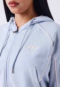 Lichtblauwe hoodie gemaakt van zachte stof, met een ritssluiting, contrasterende witte stiksel en een klein logo op de borst.