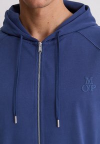 Mann trägt einen blauen Kapuzenzipper mit einem kleinen geprägten "M O P"-Logo auf der linken Brust.