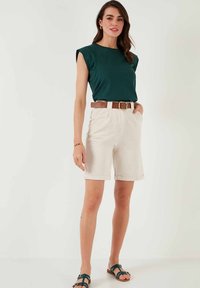 Top verde a maniche corte, pantaloni beige a vita alta con tasche e cintura marrone. Sandali neri con cinturini. Design minimalista, stile casual.