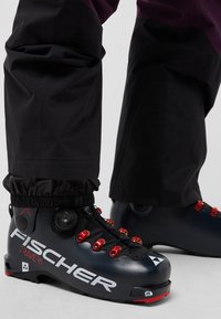 Stivali da sci neri con hardware e lacci rossi, caratterizzati da una finitura opaca. Abbinati a pantaloni impermeabili neri a livello della caviglia.