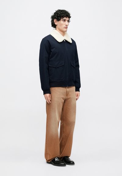 Temno modra jakna z zadrgo, kremnim ovratnikom iz umetnega krzna, dvema sprednjima žepoma in rebrastimi manšetami. Kombinirana s svetlo rjavimi širokimi hlačami in črnimi čevlji.