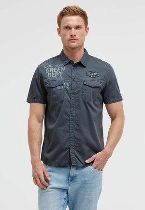 Mann trägt ein dunkles kurzärmeliges Hemd mit Knöpfen und bedrucktem Text sowie hellblaue Jeans, steht mit einer Hand in der Tasche.