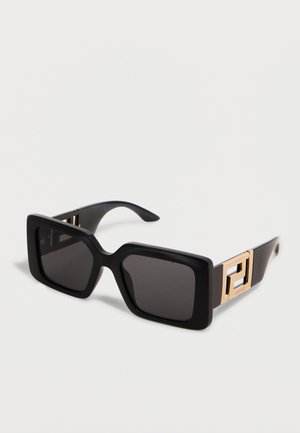 Lunettes de soleil rectangulaires noires avec des montures épaisses et des accents du logo Versace doré sur les branches, présentées sur un fond clair uni.