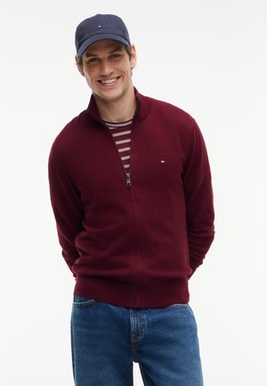 Lächelnder Mann mit einer marineblauen Kappe, einem bordeauxfarbenen Reißverschluss-Pullover über einem gestreiften Hemd und blauen Jeans, der vor einem schlichten weißen Hintergrund steht.