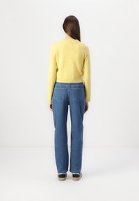 Suéter amarelo de tricot com bainha canelada, combinado com jeans de cintura alta azuis com bolsos traseiros. Modelo em pé contra um fundo liso.