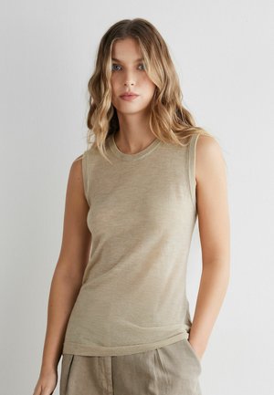 Frau mit welligem blondem Haar, die ein ärmelloses beiges Oberteil und beige Hosen trägt, vor einem schlichten hellgrauen Hintergrund stehend.