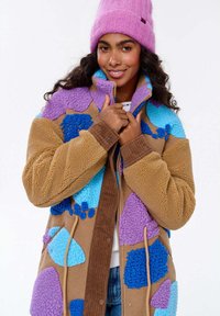 Kobieta uśmiechająca się i trzymająca kołnierz kolorowej kurtki z patchworku z polaru, nosząca różową dzianą czapkę i niebieskie dżinsy.