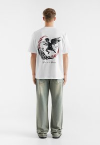 White cotton t-shirt features a black and red graphic design on the back, depicting an angel with text "Love is a Dream." Casual fit.