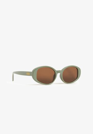 Lunettes de soleil vert olive avec des montures ovales surdimensionnées et des verres teintés bruns. Présente le logo Vans sur le côté.