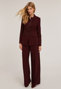 Burgundowy garnitur z dopasowaną marynarką z dwiema przednimi kieszeniami oraz szerokimi spodniami. Gładki materiał o eleganckim wykończeniu.