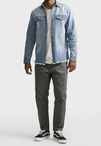 Man in een lichtblauwe versleten denimjas over een wit T-shirt, olijfgroene broek en zwart-witte sneakers, staande tegen een effen achtergrond.