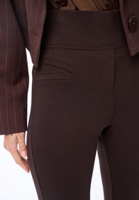 Bruine aangesloten broek met een hoge tailleband, voorzien van zijzakken en een gladde textuur. De close-up toont een bruin blazer met een streepdessin.