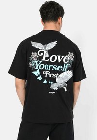 Zwarte T-shirt met een grafisch ontwerp waarop de tekst "Heb Eerst Houd Van Jezelf" staat, met vogels, rozen en vlinders in wit en lichtblauw.