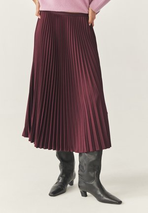Woman wearing a purple pleated midi skirt, pink sweater, and black heeled leather boots, standing with hands on hips.