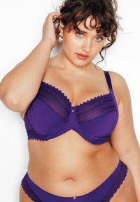Soutien-gorge en dentelle violette avec un panneau texturé, un soutien sous-armature, des bords décoratifs festonnés et un détail de bouton sur le devant. Culottes assorties violettes.