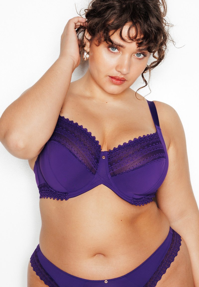 Soutien-gorge en dentelle violette avec un panneau texturé, un soutien sous-armature, des bords décoratifs festonnés et un détail de bouton sur le devant. Culottes assorties violettes.