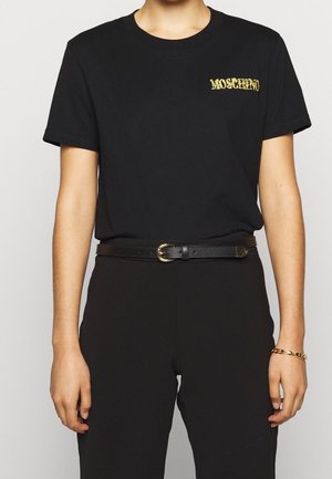Person trägt ein schwarzes Moschino-T-Shirt mit goldenem Logo, schwarze Hose, schwarzen Gürtel mit goldener Schnalle und ein goldenes Kettenarmband am rechten Handgelenk.