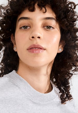 Ung kvinde med krøllet mørkt hår iført diamantørestikker og en lysegrå rundhalset skjorte, kigger let nedad.