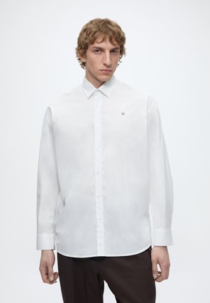 Junger Mann mit lockigem blondem Haar, trägt ein weißes Hemd mit Knöpfen und dunkle Hose, steht vor einem einfarbigen weißen Hintergrund.