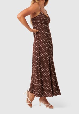 Mujer vestida con un vestido maxi marrón sin mangas con lunares y tirantes finos, y sandalias beige de tacón, de pie con las manos en la cintura.