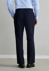 Pantalon bleu marine à coupe droite, doté de deux poches arrière avec fermetures à boutons. Fabriqué en tissu lisse, associé à des chaussures noires.